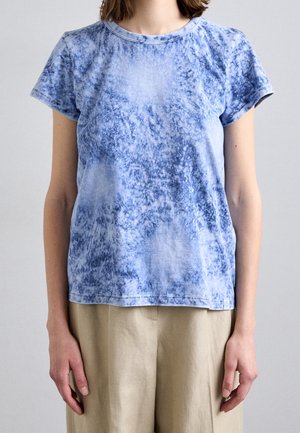 Personne portant un t-shirt à manches courtes bleu tie-dye et un pantalon beige, debout devant un fond gris clair uni.