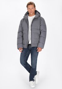 Grå polstret jakke med hætte, lynlås og elastiske manchetter. Parret med mørke jeans og hvide sneakers, stående foran en ensfarvet baggrund.