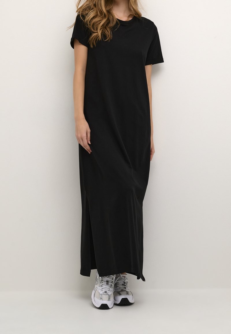 Schwarzes Maxikleid mit kurzen Ärmeln, lässiger Passform und einem seitlichen Schlitz. Getragen mit weißen und grauen gemusterten Sneakers. Glatte Baumwolltextur.