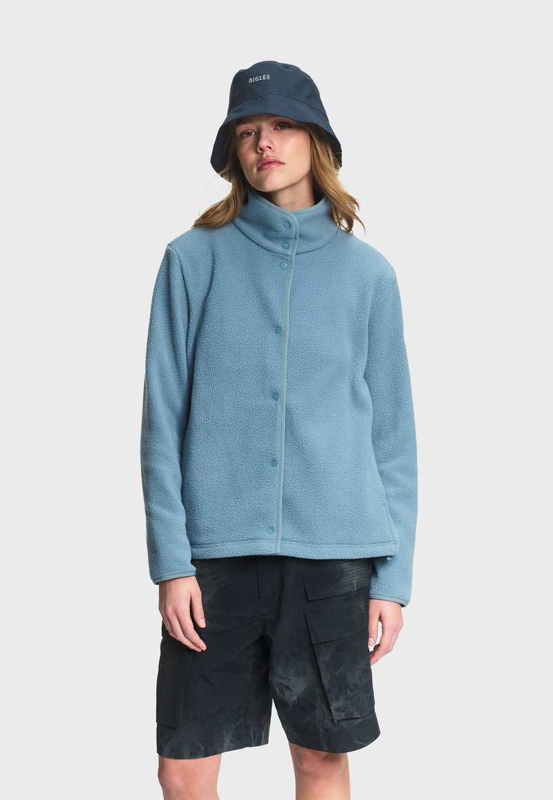 Femme portant une veste polaire bleu clair à col montant, un short cargo noir et un chapeau bob bleu marine avec le logo "AIGLE".