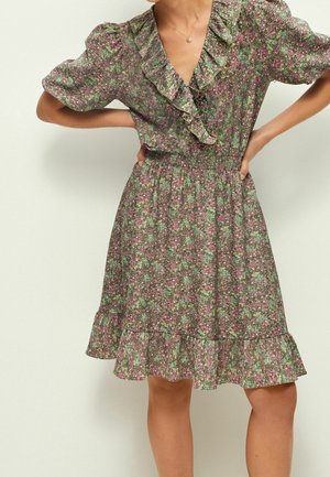 Femme portant une robe verte fleurie à volants avec manches bouffantes et décolleté en V, mains sur les hanches, debout devant un fond uni.