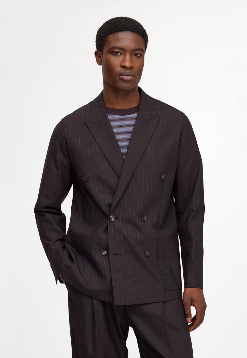 Man wearing a dark pinstripe double-breasted suit jacket and matching trousers with a striped shirt underneath, standing against a plain background.