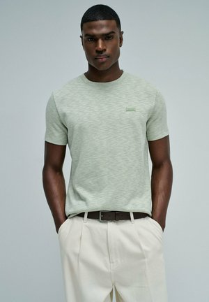 Hombre con las manos en los bolsillos, vestido con una camisa de manga corta verde claro y pantalones crema con cinturón oscuro sobre un fondo gris liso.