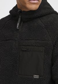 Svart fleece-pullover med høy krage, halv glidelås foran og en kontrastfarge lomme; har et strukturert, fluffy stoff og logo-patch.