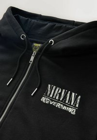 Juodas užtrauktukas su kapucėle, ant krūtinės užrašyta "NIRVANA NEVERMIND" baltomis raidėmis, o vidinėje apykaklėje pavaizduota grupės šypsenėlių logotipas.