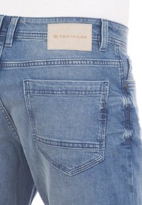 Vaqueros de mezclilla azul claro con una etiqueta de cuero que dice "TOM TAILOR", con un bolsillo trasero y detalles de costura visibles.