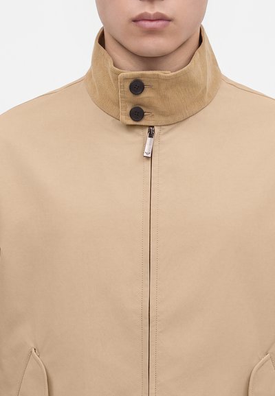 Homme portant une veste beige avec un col en velours côtelé haut fermé par deux boutons noirs et une fermeture éclair centrale.