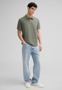 Olijfgroen katoenen polo shirt met een kraag en drie knopen, gecombineerd met lichtblauwe loszittende spijkerbroek en witte sneakers.
