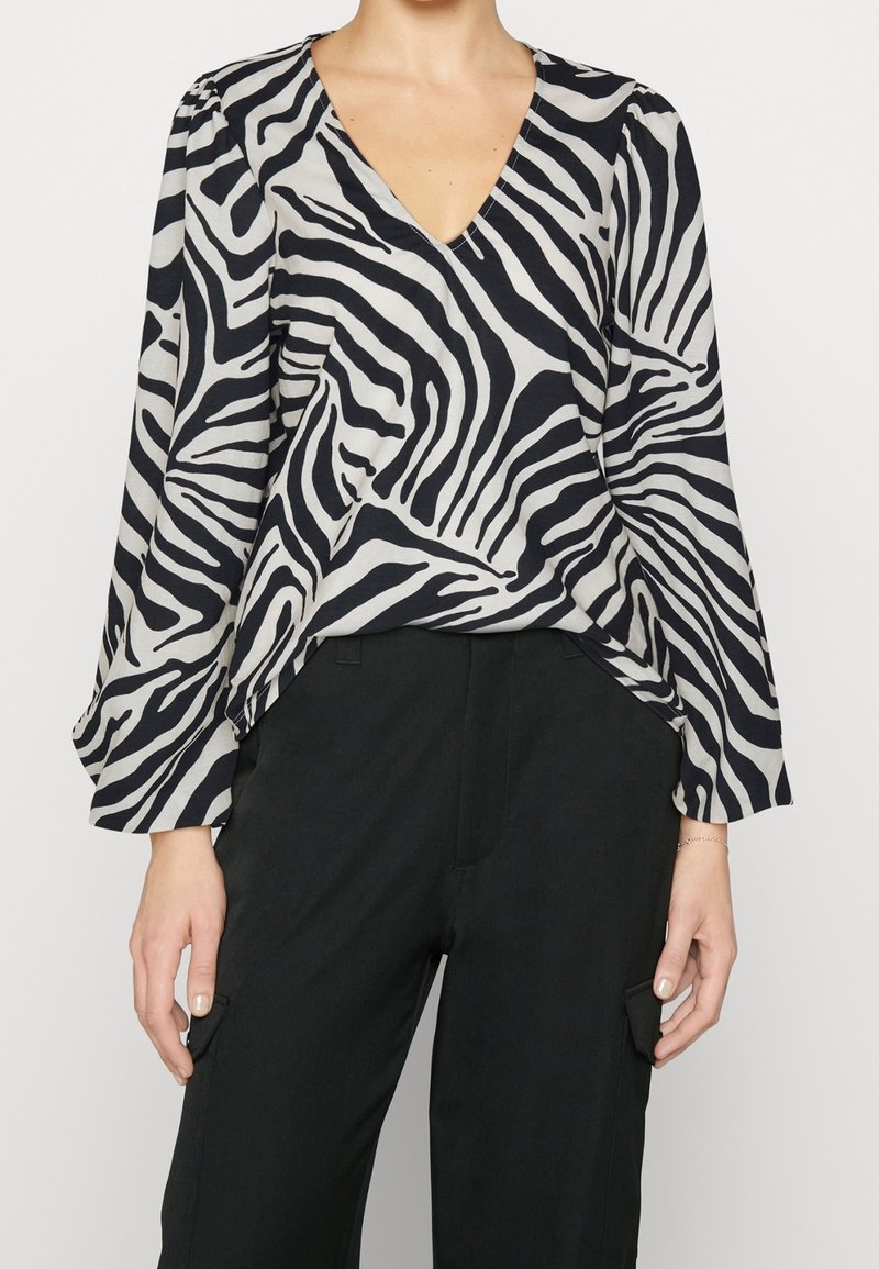 Blouse à imprimé zèbre noir et blanc avec un col en V, des manches longues et une coupe décontractée, associée à un pantalon noir avec des poches latérales.