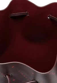 Okrągła torba-bucket w ciemnoczerwonym skórzanym wykończeniu, z falistymi krawędziami i plecionymi uchwytami. Wnętrze zdobi subtelny wzór logo.