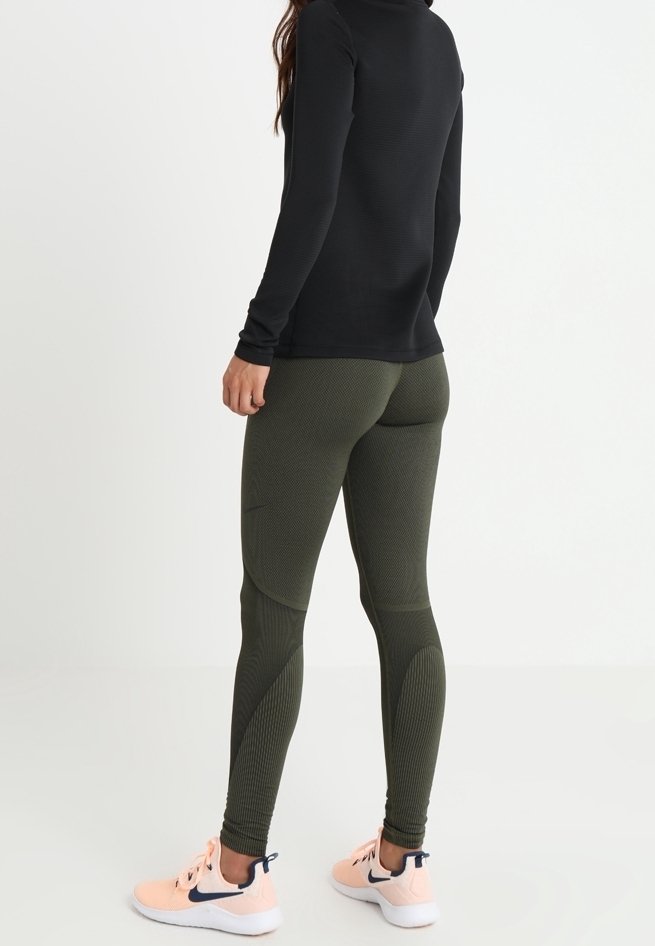 Maglia nera a maniche lunghe abbinata a leggings verdi oliva dalla textures, con design aderente e sneakers arancioni con accenti scuri.