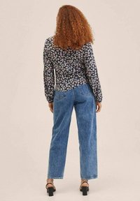 Blumenmuster-Bluse mit langen Ärmeln auf schwarzem Hintergrund, kombiniert mit hochtaillierten blauen Jeans. Zu dem Outfit passen die Riemchensandalen.