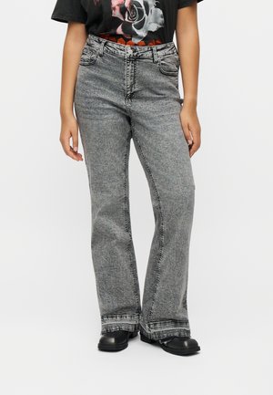 Persona con vaqueros grises de tiro alto y corte holgado, camiseta negra con un diseño gráfico de una rosa y zapatos negros, de pie frente a un fondo liso.