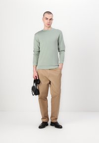 Camisa de manga longa verde claro combinada com calças bege. Pequena bolsa preta segurada em uma das mãos, usada com botas pretas, contra um fundo simples.