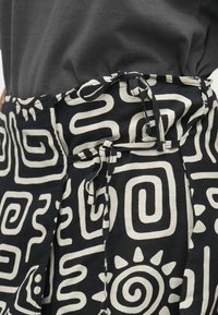Black fabric with white abstract patterns, featuring a tied waist and side pockets. The design includes spirals and geometric shapes.