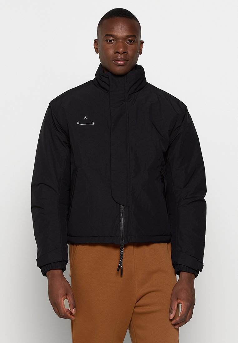 Chaqueta negra corta con un cuello alto, que presenta un cierre de cremallera y un detalle de logotipo minimalista en el pecho. Textura de tela suave y lisa.