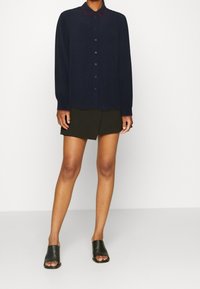 Marineblaues Langarmhemd mit rotem Kragenbesatz, Knopfleiste, kombiniert mit einem schwarzen Minirock und schwarzen offenen Riemchensandalen.