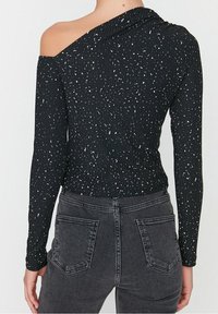 Svart ribbad topp med design för ena axeln, prydd med vita prickar. Kombinerad med högmidjade grå jeans med bakfickor.