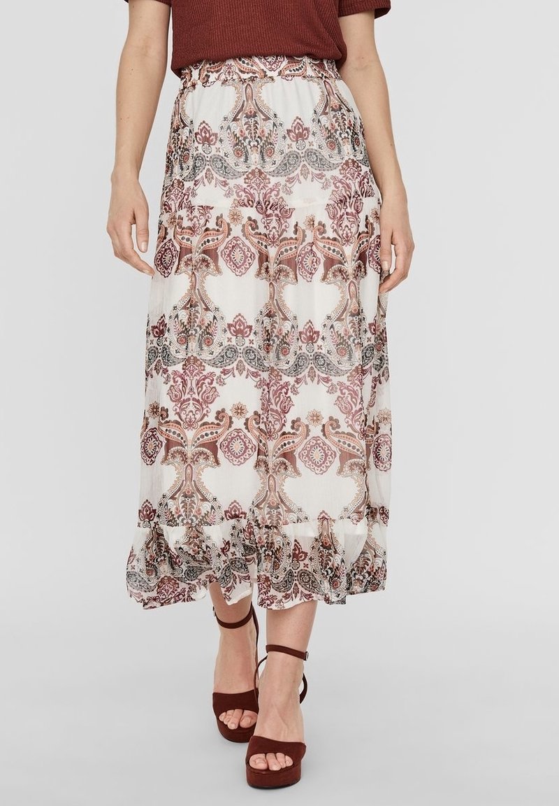 Femme portant une jupe midi blanche à motifs cachemire rouges et gris, un haut couleur rouille, et des sandales à talons hauts assorties couleur rouille.