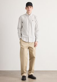 Gestreepte button-up shirt in wit en grijs, gecombineerd met beige broeken. Een zwarte pet en donkere klompen maken de outfit compleet. Minimalistisch ontwerp.