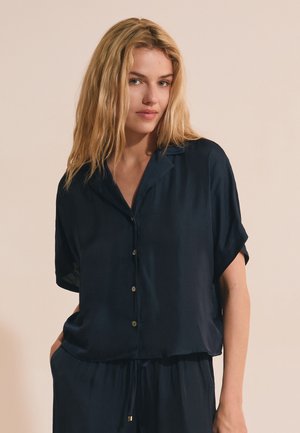 Femme blonde aux cheveux lâchés, portant une chemise bleu marine à manches courtes boutonnée et un pantalon assorti sur fond beige.