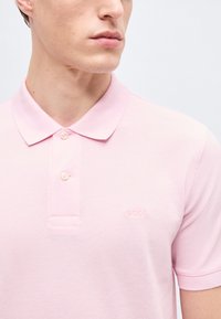 Νεαρός άνδρας που φοράει polo μπλουζάκι BOSS ανοιχτό ροζ με δύο κουμπιά κουμπωμένα, κοιτάζει προς τα πλάγια σε απλό φόντο.