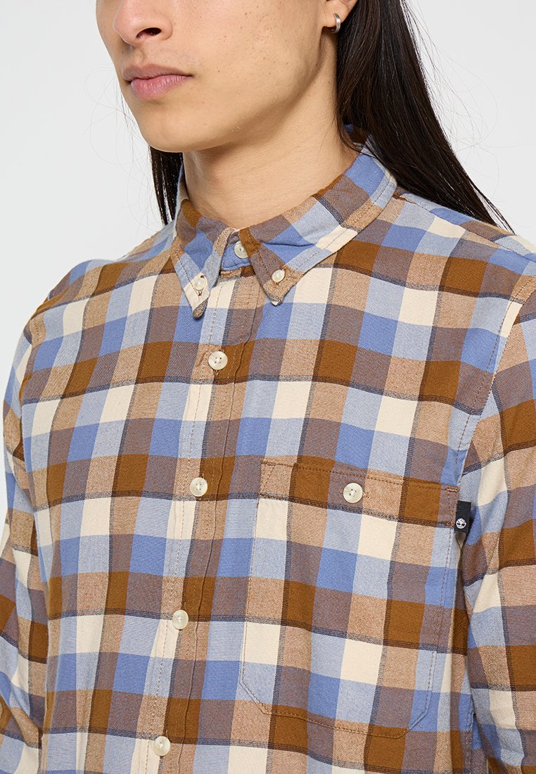 Camicia a quadri marrone, blu e crema con colletto, due tasche sul petto e bottoni bianchi. Tessuto leggero e strutturato.