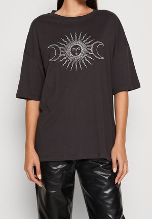 Osoba nosząca czarną, oversize koszulkę z białą grafiką słońca i półksiężyca z przodu, zestawioną z błyszczącymi czarnymi spodniami.