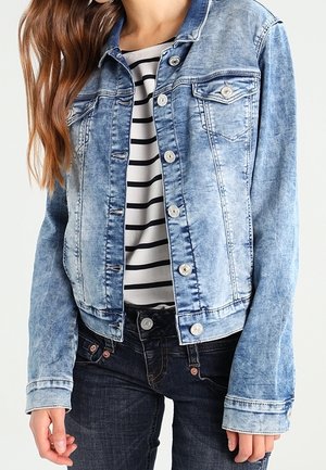 Vaaleansininen farkkutakki, jossa nappikiinnitys edessä, päällä valko-musta raidallinen paita ja tummat farkut.