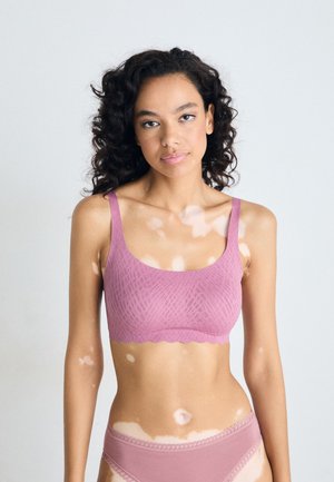 Young woman with curly hair and vitiligo wearing a pink textured bra and matching underwear against a plain light background.