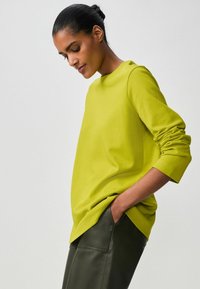 Limoengroene lange mouw shirt met een ronde halslijn en opgerolde manchetten, gedragen met zwarte leren hoge taille broek met een gladde textuur.