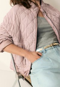 Quiltet bomberjakke i lys pink, med ribbet krave, lynlåslukning og sidelommer, parret med en grå tanktop og lyseblå jeans.