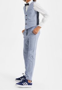 Un garçon portant un gilet bleu clair et un pantalon avec une chemise à pois blanche et des baskets bleu marine, les mains dans les poches, se tenant de profil.