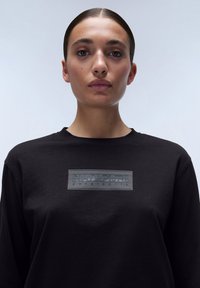 Zwarte longsleeve t-shirt gemaakt van katoen, met een gestructureerde grijze logo patch op de borst. Eenvoudig ontwerp met een ronde halslijn.