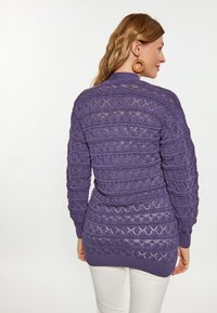 Pulover tricotat violet cu textură patterned, mâneci lungi și croială lejeră, purtat cu pantaloni albi.