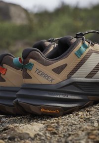 Ζευγάρι καφέ και μπεζ παπουτσιών τρεξίματος Terrex με μαύρες σόλες και πέλμα Continental, τοποθετημένα σε πετρώδες έδαφος σε εξωτερικό χώρο.
