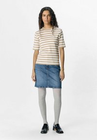 Randig beige och vit kortärmad topp, blå denimkjol, grå ribbade strumpbyxor, svarta lackskor, står mot en neutral bakgrund.