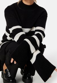 Übergroßer schwarz-weiß gestreifter Pullover mit hohem Kragen, gerippter Textur und weiten Ärmeln. Kombiniert mit schwarzen Hosen und klobigen Schuhen.