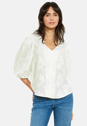 Sieviete, tērpusies baltā ziedu tekstūru blūzē ar pūderpiedurknēm un zilās džinsās, stāvot ar vienu roku aiz muguras.