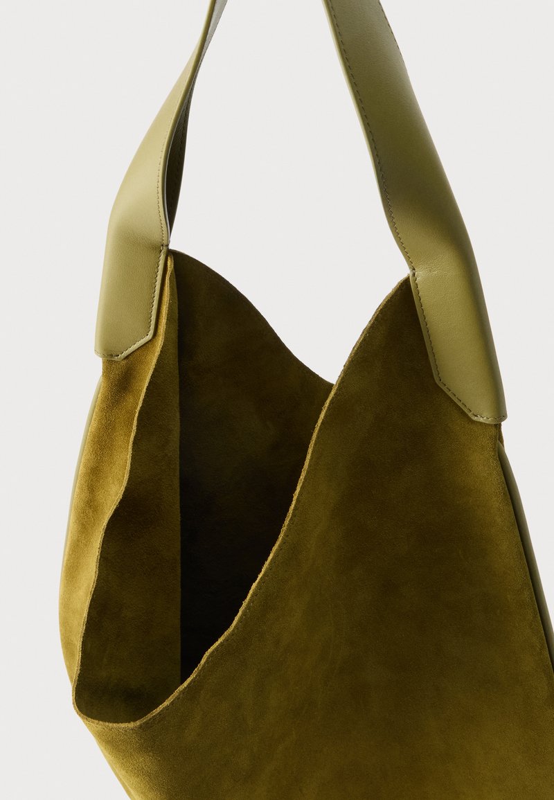 Sac d'épaule en daim vert olive doux avec une large sangle en cuir et un haut ouvert dévoilant un intérieur spacieux.