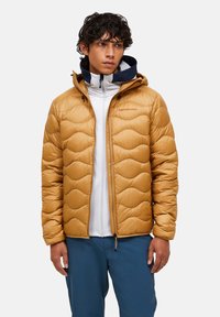 Ung mann iført en brun quiltesjakk over en hvit zip-up hettegenser og blå bukser, står mot en enkel lys bakgrunn.