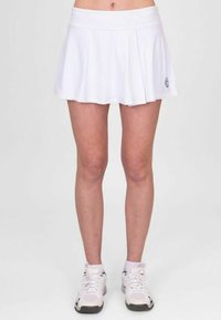Falda plisada blanca con una textura suave, que presenta una cinturilla ancha y un pequeño detalle de logo. Combinada con zapatillas deportivas blancas.