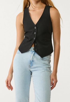Vrouw die een mouwloze zwarte gilet met knopen draagt, gecombineerd met lichtblauwe jeans met hoge taille, staand tegen een effen witte achtergrond.