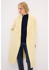 Abrigo amarillo de doble botonadura con una textura suave, grandes botones amarillos y una silueta larga y recta, llevado sobre un suéter y pantalones azul marino.