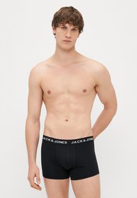 Črne bokserice z mehkim pasom, ki vsebujejo bele oznake "JACK & JONES". Tkanina se zdi gladka in prilegajoča. Model prikazuje nevtralen položaj.