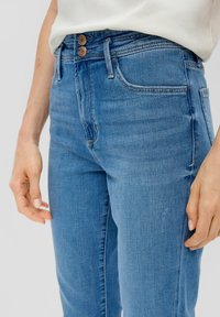 Jean en denim bleu taille haute avec deux boutons, design à cinq poches et texture lisse, présentant des marques d'usure subtiles sur le devant.