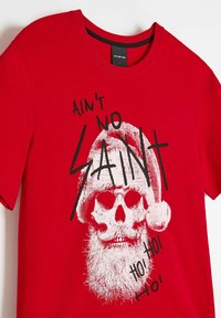 Czerwona bawełniana koszulka z grafiką czaszki w czapce i brodzie Mikołaja. Napis w czerni: "AIN'T NO SAINT HO! HO! HO!"