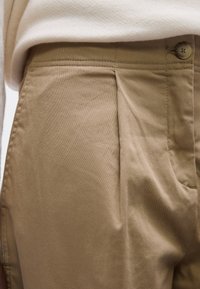 Bézs pamut nadrág redőkkel, lapos elejű, gombos záródású, oldalzsebbel, sima textúrájú, derékszinten, világos háttér előtt bemutatva.