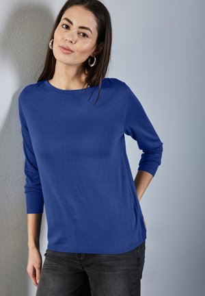 Haut à manches longues bleu en tissu tricoté lisse avec un col rond et des poignets côtelés, porté avec un jean foncé sur un fond neutre.