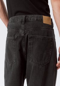 Jeans de mezclilla negra con textura desvaída, que cuentan con dos bolsillos traseros, una etiqueta de marca de cuero marrón y un diseño de cinturilla tradicional.